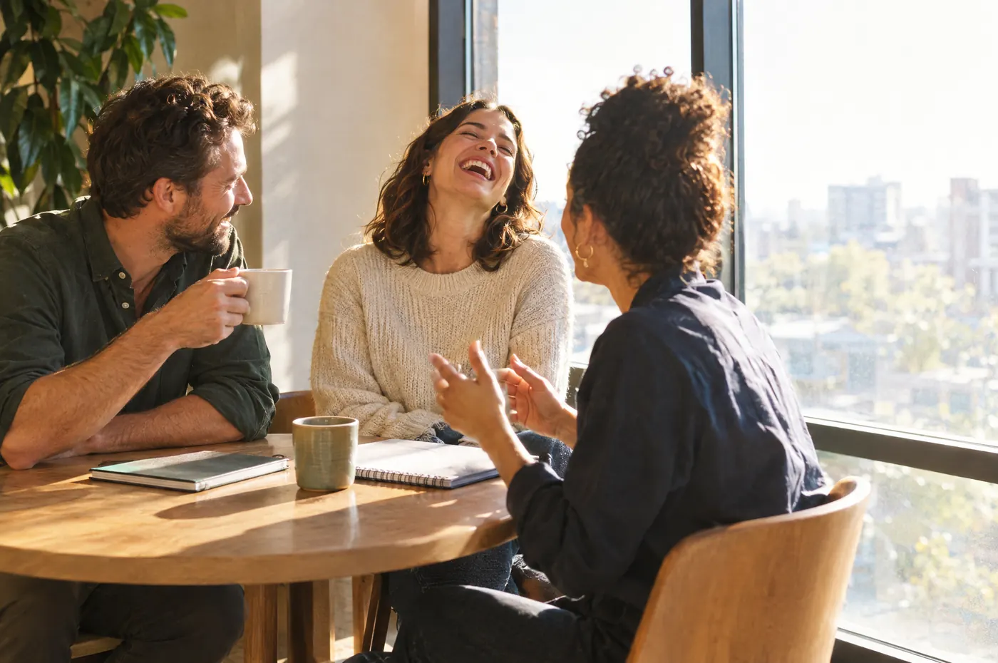 Three people laughing in conversation around a round table by a window.